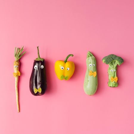 Various vegetables with googly eyes and bow tie pasta on pink background