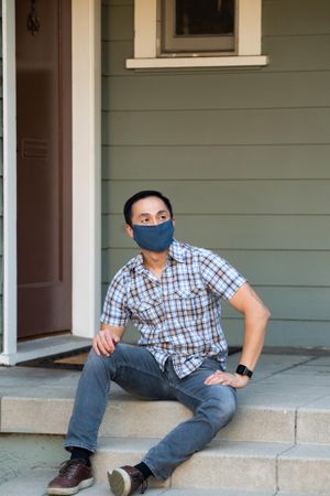 Man wearing PPE mask in casual clothing sitting on porch smiling and looking away