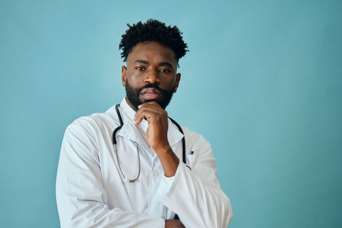 Thoughtful Black male doctor in blue studio with hand to his chin