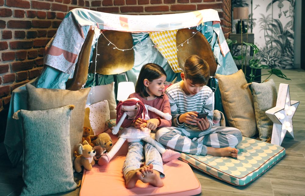 Happy little girl embracing rag doll while looking boy playing video game with phone sitting on tent