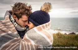 Closeup of young couple embracing under blanket in a cold day with ocean and dark cloudy sky in the background 5RWnB4