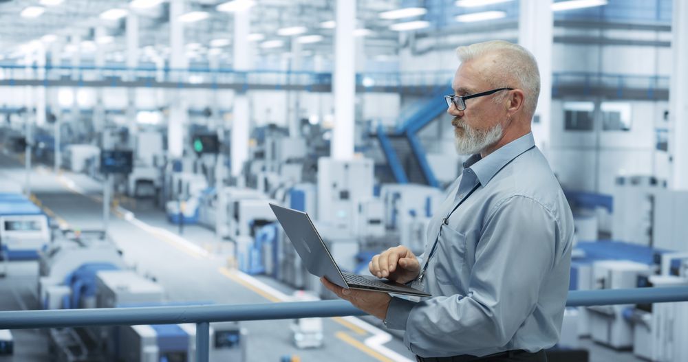 Portrait of a Handsome Mature Engineer with Glasses Using Laptop Computer and Looking Around a Factory Facility with Equipment Producing Modern Electronic Components for Different Industries