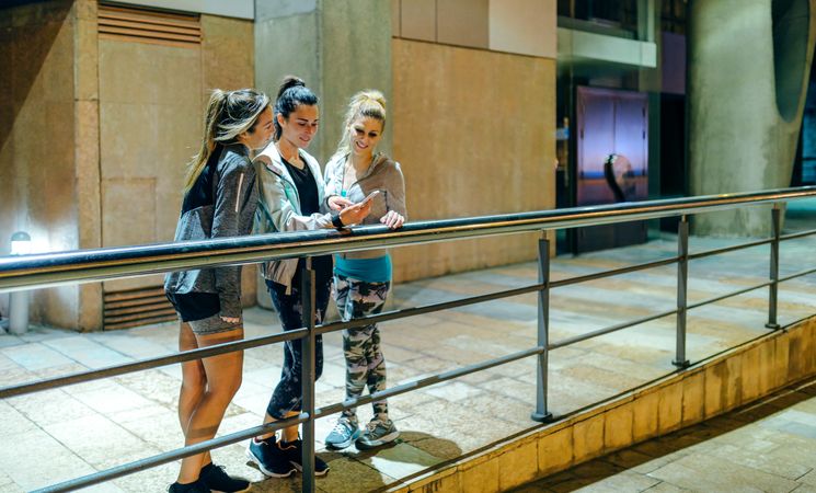 Smiling sporty women looking at cellphone while leaning on banister after training at night
