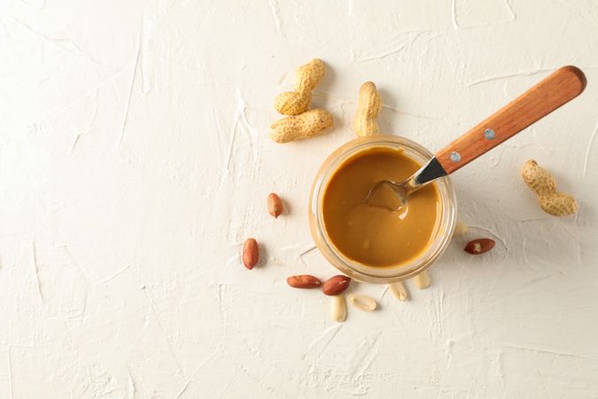 Glass jar with creamy peanut butter and spoon, and peanut on background, space for text and top view