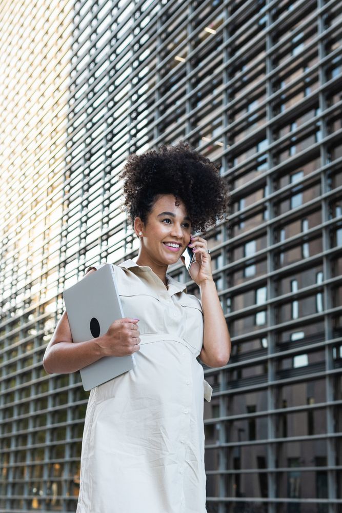 Smiling Black pregnant woman talking on smartphone and carrying a laptop