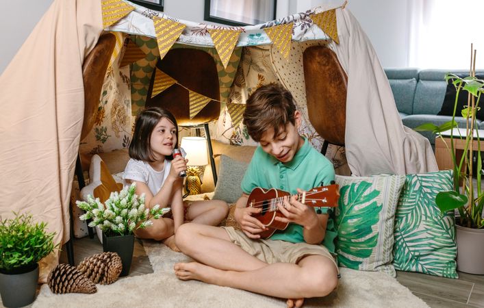 Happy children playing a ukulele and singing on handmade shelter tent in living room at home