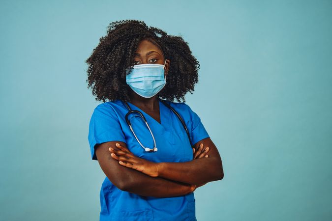 Black female doctor standing with arms crossed with face mask and stethoscope in blue scrubs