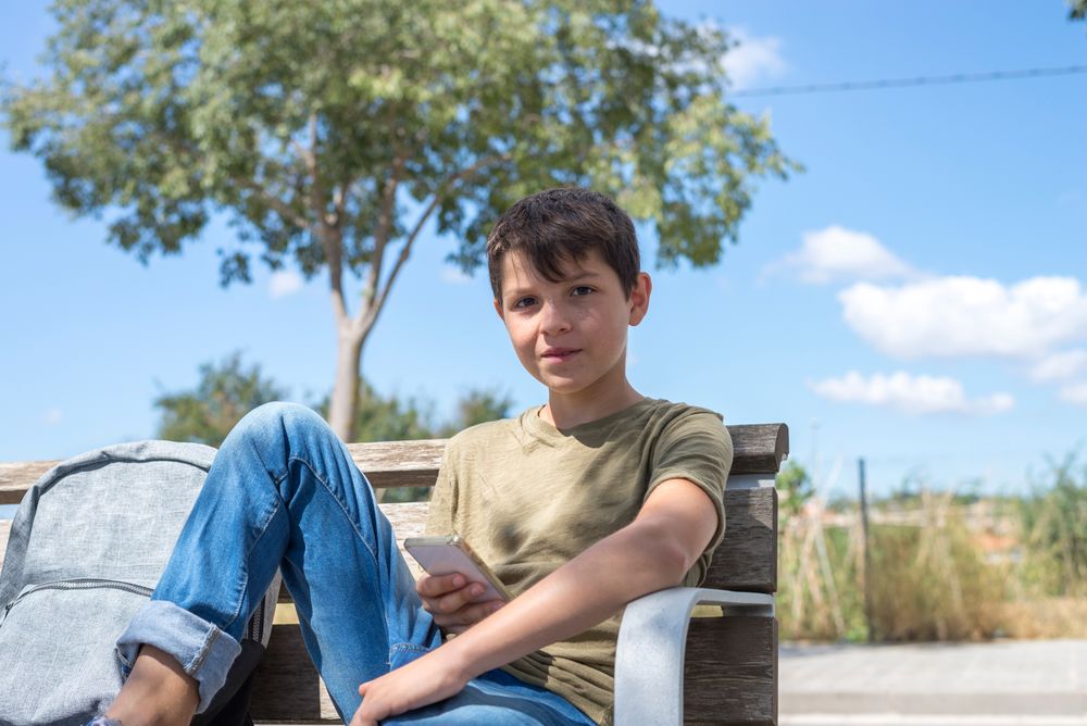 Boy with backpack sitting outside holding smartphone