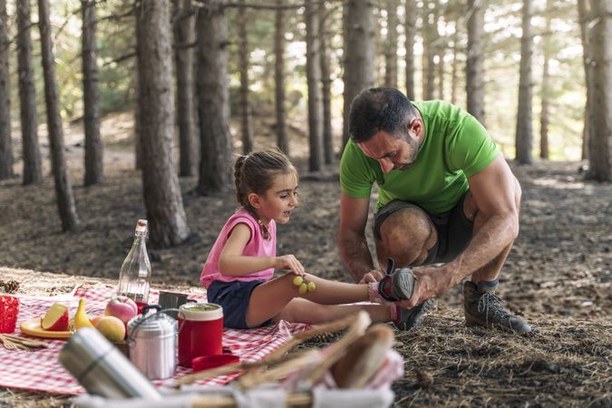 Caring father helping his young daughter with her shoe during a family picnic in the forest.