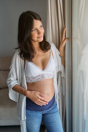 Content woman with bared pregnant belly standing against window