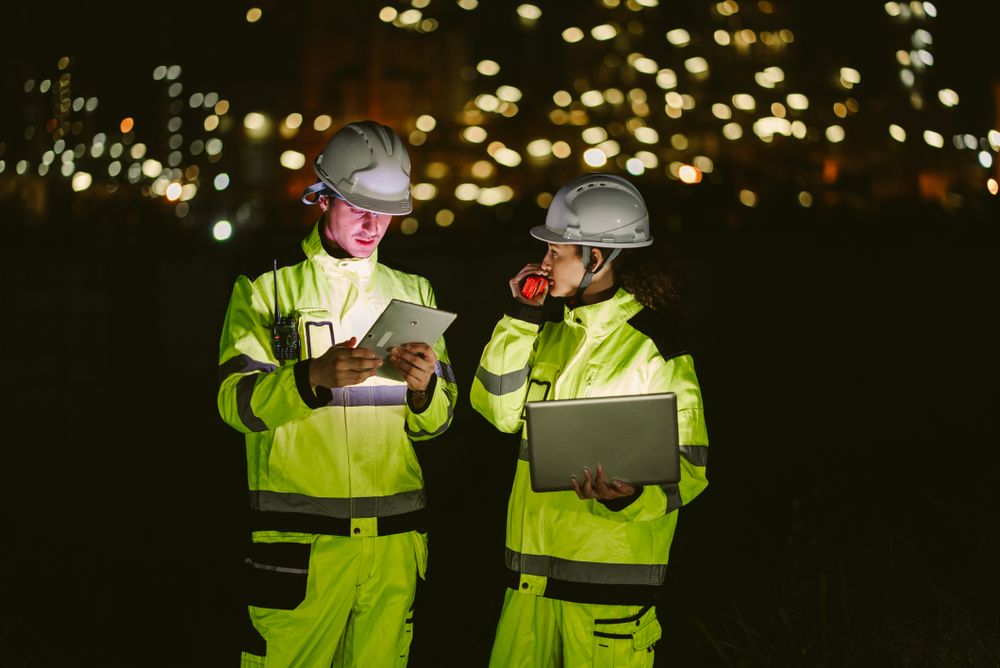 Team of two industrial workers using laptop, walkie-talkie, and phone at night