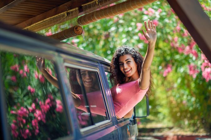 Happy female in pink t-shirt leaning out of car window and waving