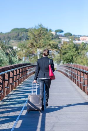 Back of woman walking confidently across bridge with roller suitcase