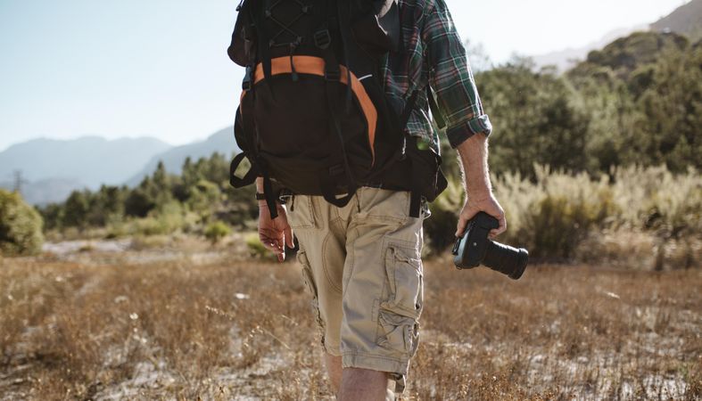 Rear view of a older man carrying a backpack hiking in nature holding a digital camera