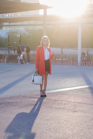 Blonde businesswoman wearing red jacket and standing in the city park while looking to camera