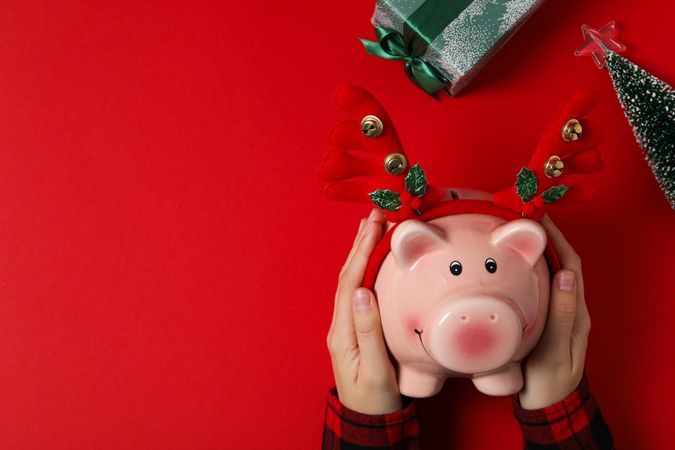 Piggy bank with Christmas decor in hands and gift box on red background, space for text
