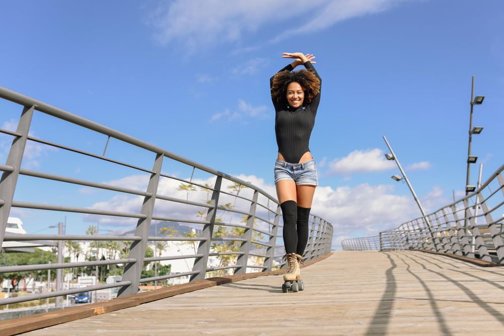 Woman with afro hairstyle in roller skates on wooden bridge with arms above head