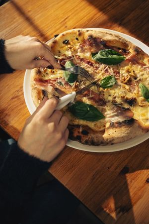 Pizza with basil being cut with knife and fork on a plate at table