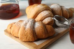 Tasty croissants and cups with tea on wooden cutting board, close up