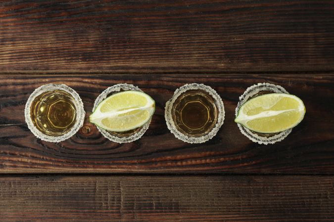 Tequila shots with salt and lime slices on wood table, top view