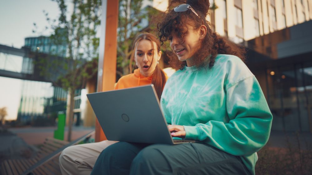 Two Young Women, One With Curly Hair In A Vibrant Tie-dye Sweatshirt, Excitedly Share A Laptop Screen Outdoors