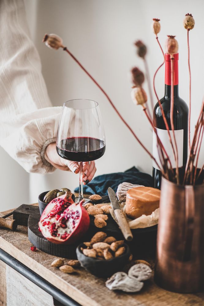 Rustic setting of women holding wine, with cheese, long candles, pomegranate, and dried poppies