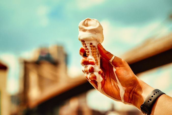 Hand holding melting ice cream cone against sky