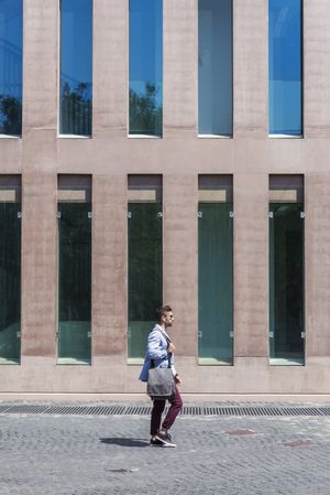 Businessman walking next to office buildings while holding a shoulder bag outdoors, vertical