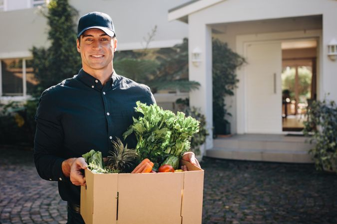 Man holding a box of fruits and vegetables outdoors looking at camera
