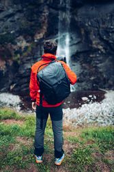 Back view of a man in red jacket with backpack standing and looking at waterfall 5oO2y5