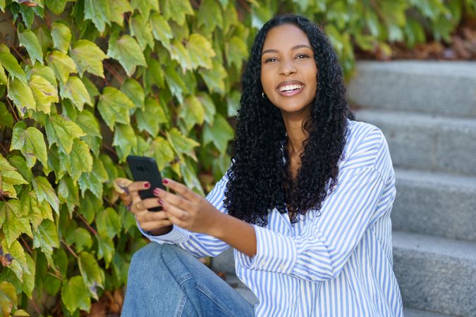 A Young Woman is Sitting on Stone Steps while Using Her Smartphone and Surrounded by Lush Greenery