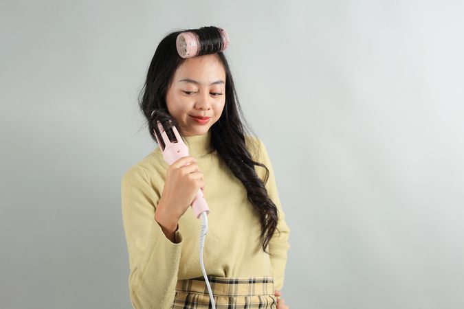 Asian Teenager using Curling Hair Iron on Grey Background.