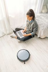 Woman working on laptop while robotic vacuum cleaner cleaning