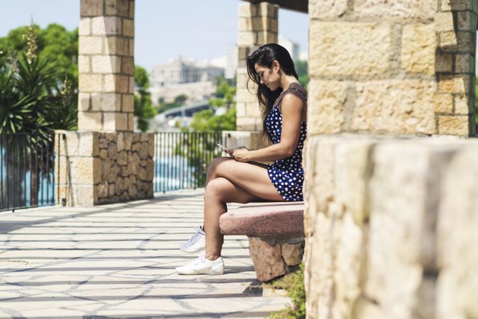 Side view of woman in blue dress sitting on a bench while using a smartphone