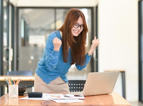 Young business woman looks down at laptop on the table with her hands up and smiles with joy