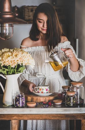 Woman in rustic kitchen pouring tea in floral cup and saucer from glass teapot