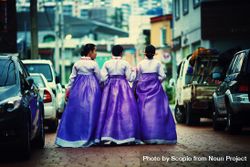 Back view of three Korean women in hanboks walking side by side between cars 0PJKg4