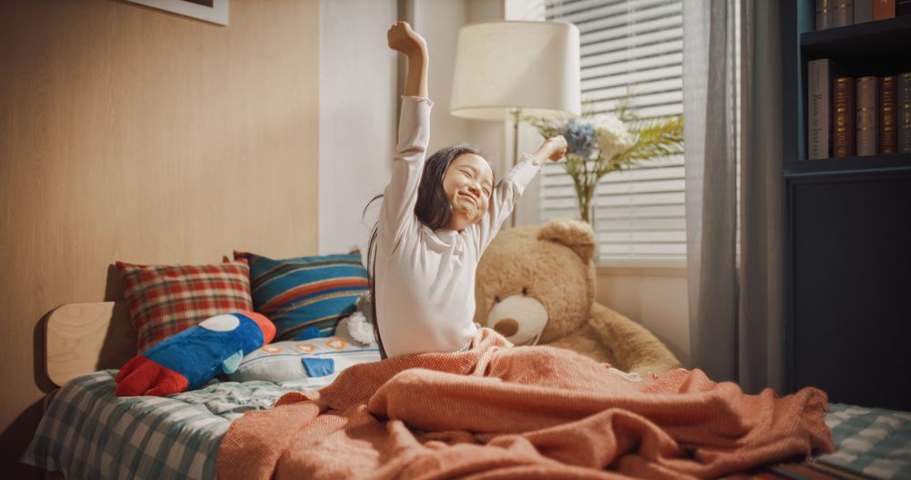 Small Kid Sitting on a Bed with Soft Toys, Stretching Her Arms, Adorably Smiling