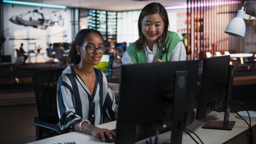 Asian Female Creative Director Giving Instructions on 3D Visuals to Black Female VFX Animator in Creative Agency Modern Office. Two Stylish Diverse Women Discussing Design on Desktop Computer