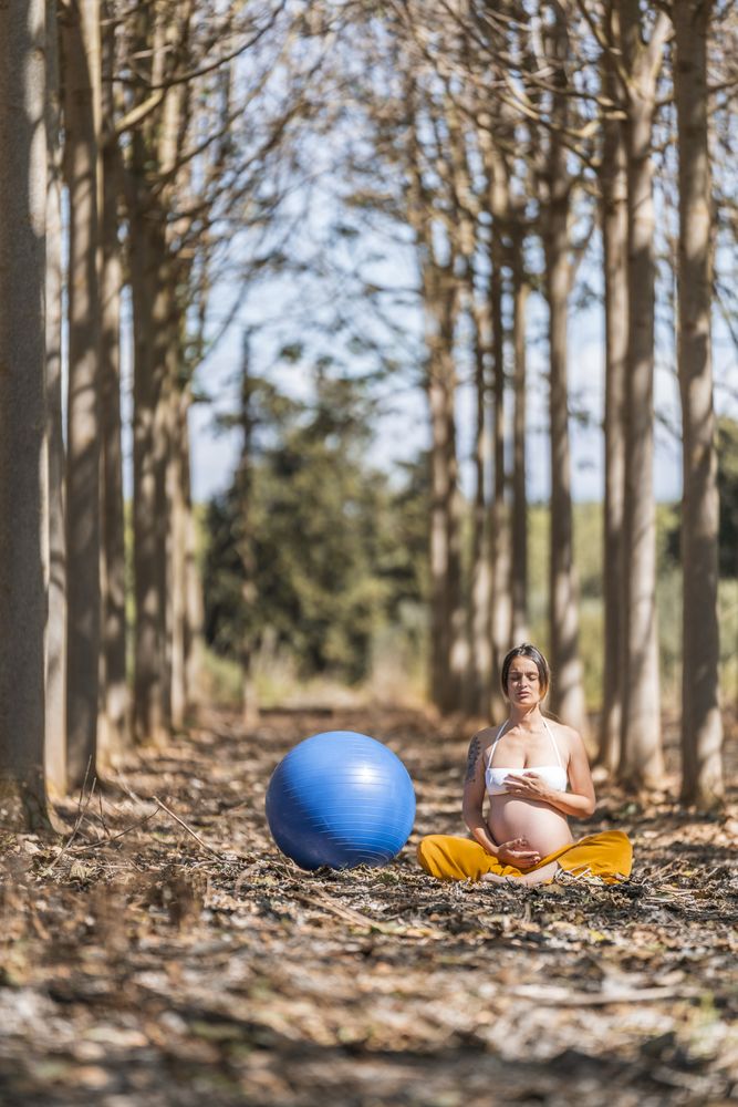 Serene adult pregnant woman meditating while sitting on ground among trees in park during sunny day