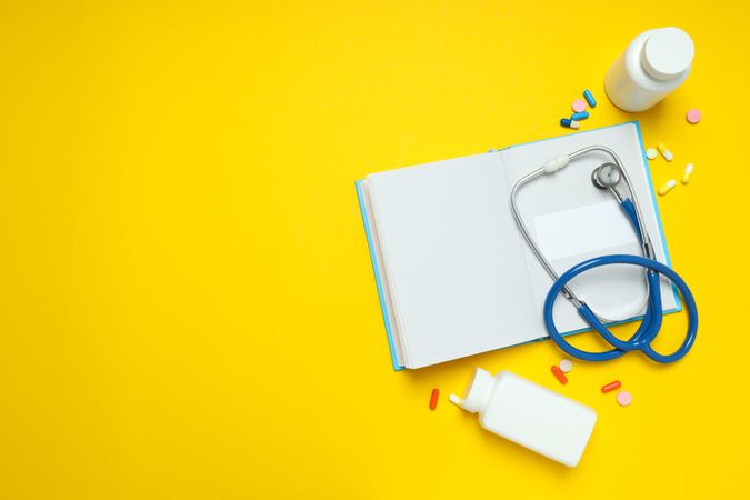 Notepad, stethoscope and jars of pills on yellow background, space for text