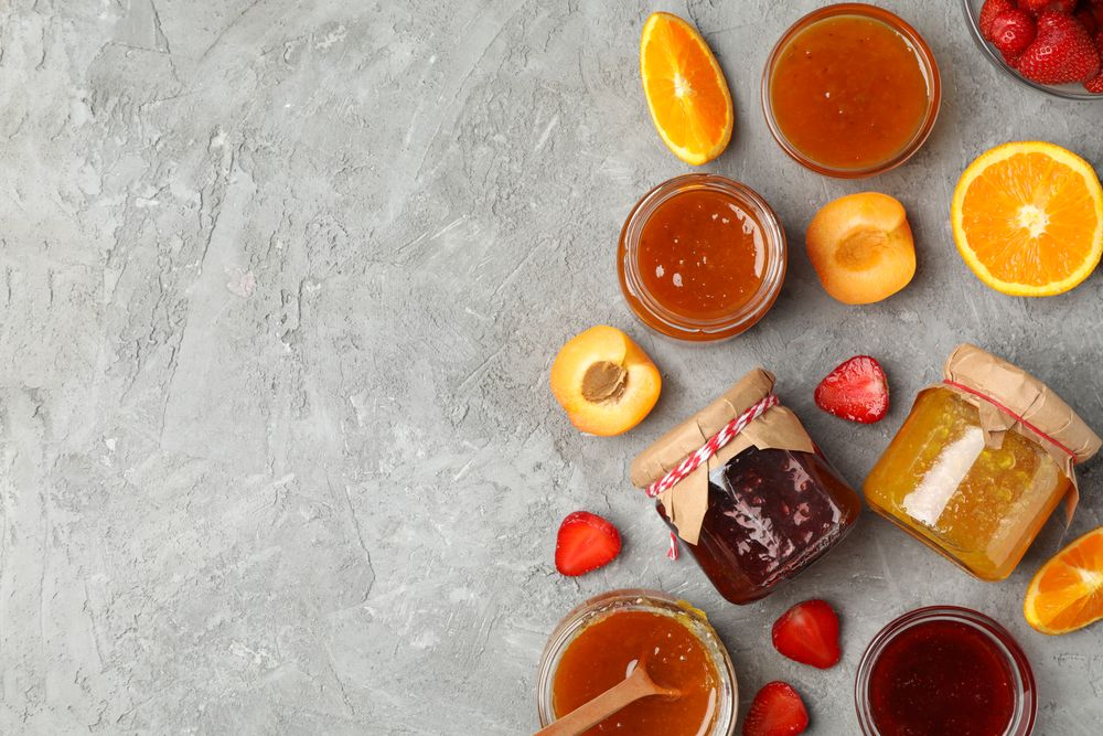 Glass jars with jam and ingredients on gray background, top view