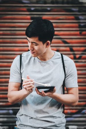 Asian man eating takeaway food with chopsticks on city street