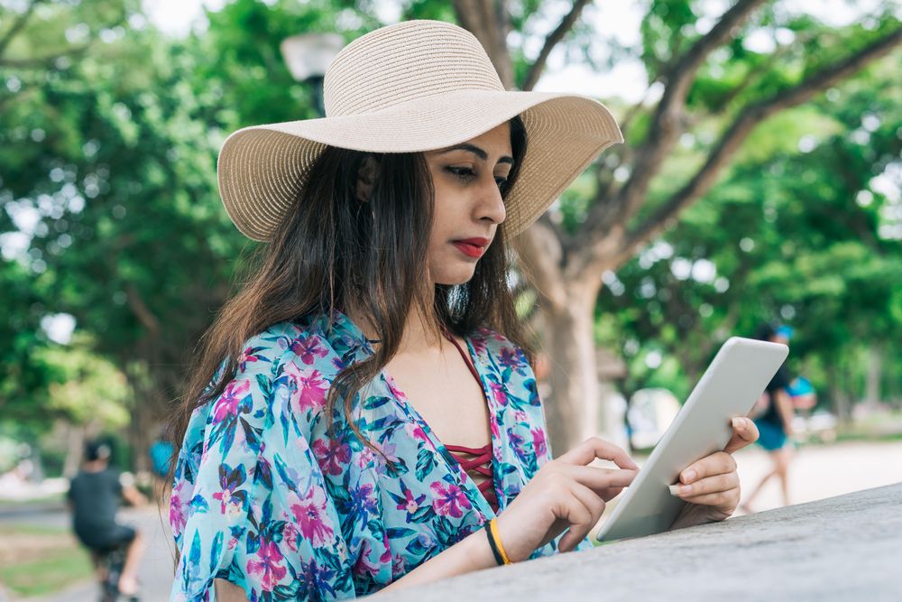 Woman wearing hat and floral dress using tablet computer outdoor