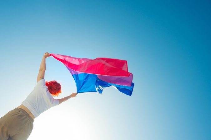 Young woman with red hair waving a bisexual Pride flag under blue sky
