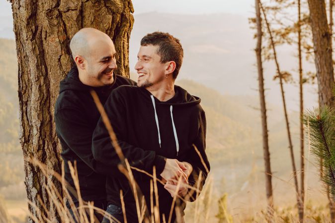 Smiling men hugging near tree in forest on hill
