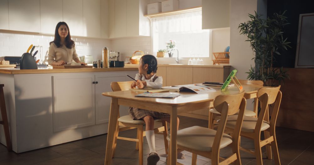 Portrait of a Korean Female Child Sitting at a Kitchen Table and Drawing While her Mother is Preparing Breakfast.
