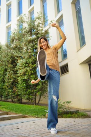 Woman in jeans and knitted vest kicking leg up outside