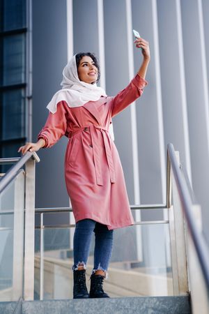 Female in headscarf and pink trench coat taking photo with her phone out, vertical composition