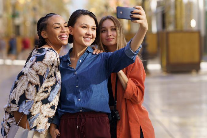 Three smiling women taking selfie with phone in city, copy space